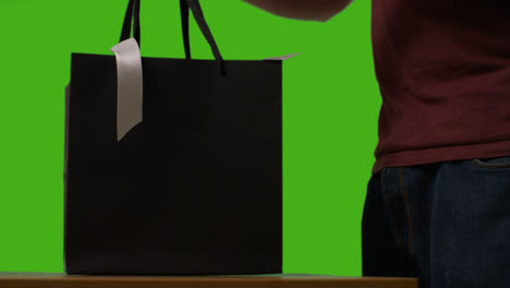 Close-Up-Of-Man-Putting-Gift-Wrapped-Present-In-Box-Into-Gift-Bag-On-Table-Shot-Against-Green-Screen